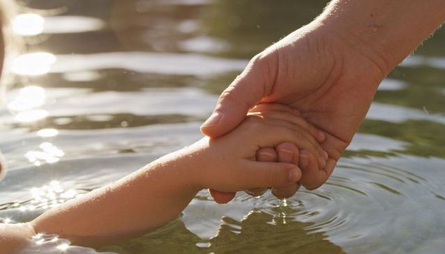 Adult hand holding child's forearm in sunlit lake water, caregiver support and bonding