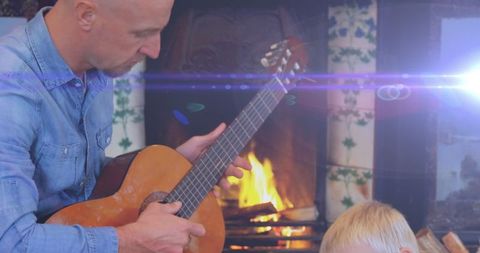 Father playing guitar near fireplace with child relaxing nearby
