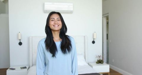 Smiling asian woman in modern minimal bedroom with smartphone, bed and wall ac
