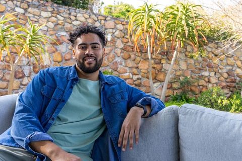 Young man relaxing on patio sofa wearing denim shirt and smartwatch in sunlit garden