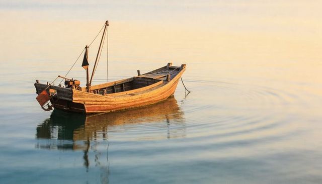 Weathered wooden boat floating on calm golden water at sunrise with soft reflection