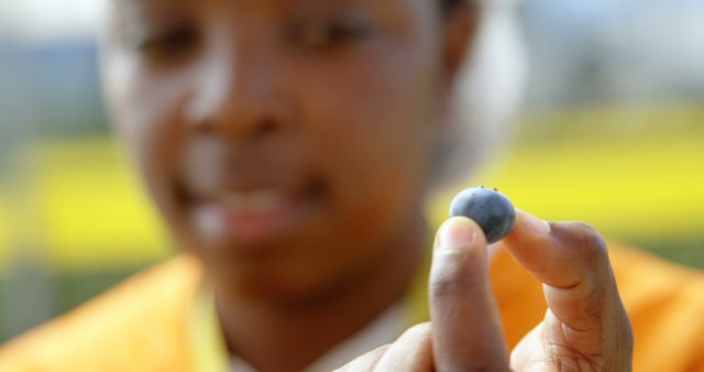 Person is holding a blueberry outdoors with a blurred background, emphasizing healthy eating and nature. Perfect for use in content related to nutrition, healthy lifestyle articles, fruit farming, or advertising fresh produce.