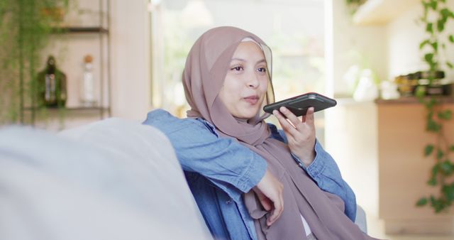 Muslim woman wearing hijab sits on couch, using voice assistant feature on smartphone. Suitable for depicting modern technology trends, communication within domestic settings, voice-activated devices, and lifestyle content. Can be used in articles or advertisements focused on technology adoption, smart home innovations, and cultural representation.