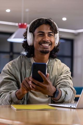 Indian man smiling holding smartphone wearing white headphones in modern coworking cafe