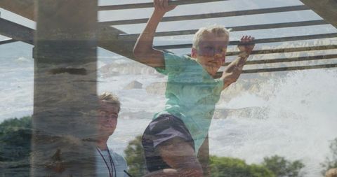 Sunlit boy climbing overhead bars with supervisor at park double exposure ocean overlay