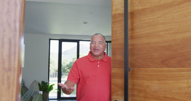 This image portrays a smiling African American man in a red shirt welcoming guests into a modern home. Ideal for use in real estate promotions, open house event flyers, or hospitality services advertising. It conveys a sense of friendliness, hospitality, and invitation.