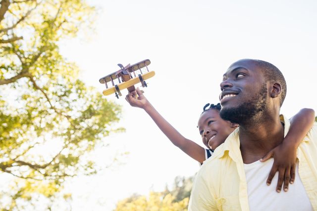 Father carrying daughter on shoulders while she holds toy airplane, enjoying sunny day in park. Perfect for themes of family bonding, outdoor activities, childhood joy, and parental love.