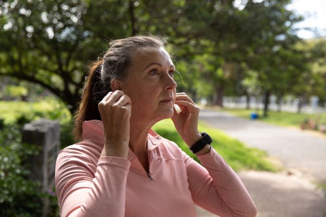 Senior woman in sports clothes putting on earphones in a park, preparing for a workout. Ideal for promoting healthy lifestyles, retirement activities, fitness routines, and outdoor exercises for mature individuals.