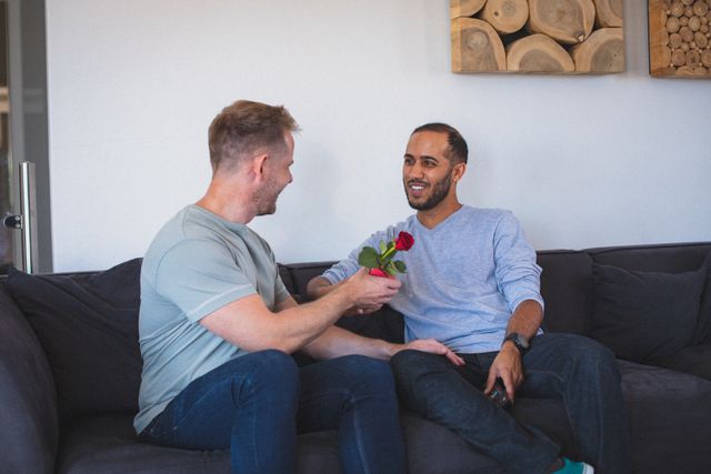 This image depicts a diverse gay couple sitting on a sofa at home, with one man giving a red rose to the other. It is an intimate and heartwarming moment, perfect for use in articles or advertisements related to LGBTQ relationships, love, and romance. It can also be used in content about staying at home during quarantine or lockdown, promoting togetherness and bonding during isolation.