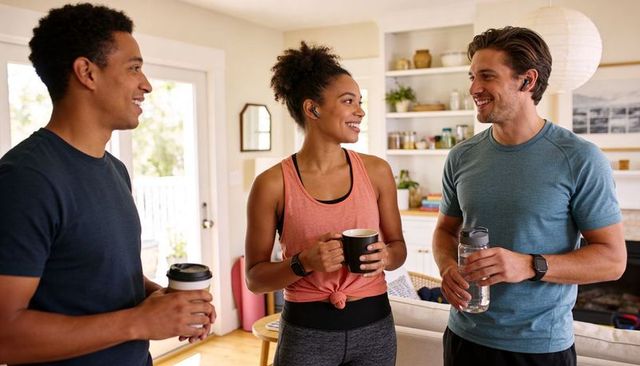 Friends chatting after workout at home, holding coffee mugs and water bottles, smiling together