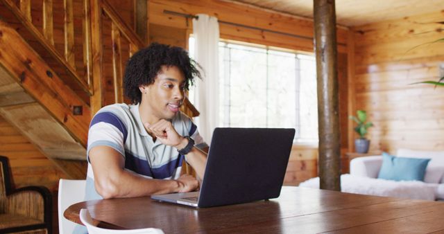 A young man is working remotely from a cozy cabin, using his laptop on a wooden table. The wooden interior and natural lighting create a comfortable and inviting workspace. This can be used for websites and articles related to remote work, freelancing, home office setups, and modern lifestyles.