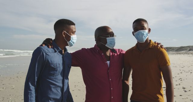 African american senior man with two adult sons in face mask embracing on beach. family time and summer holiday during covid 19 pandemic.