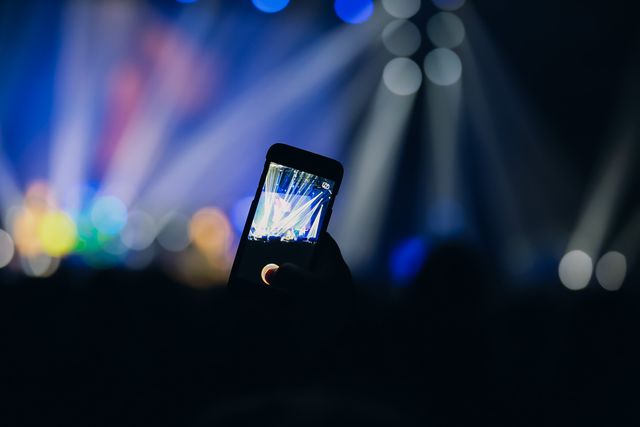 Hand holding smartphone capturing live concert moments with stage lights in background. Useful for themes like entertainment, technology, events, nightlife, and capturing memories at music festivals or concerts.