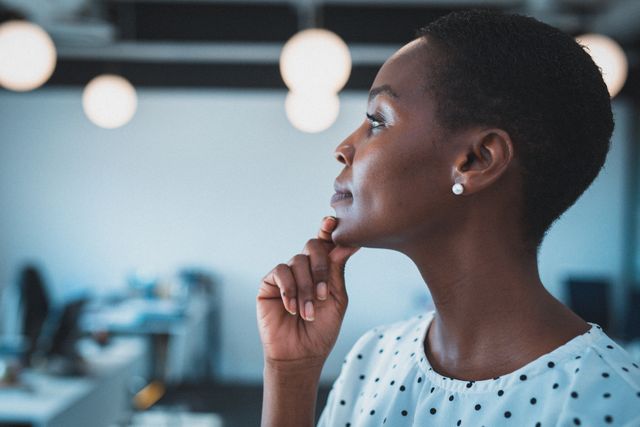 This image depicts an African American businesswoman in a modern office environment, holding her chin in thought and smiling. It can be used for business-related content, articles on workplace diversity, leadership, professional development, and corporate success stories.