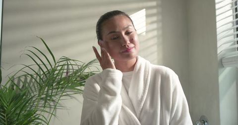 Middle-aged asian woman applying skincare in white robe at bathroom vanity with natural light