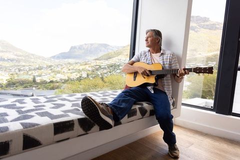 Middle-aged man playing guitar and gazing out large window over mountain valley