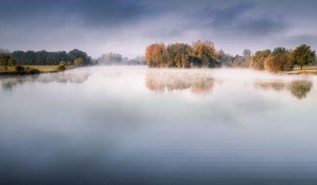 This photo captures a tranquil lake on a misty morning with autumn trees reflecting on the still water, creating a serene and peaceful atmosphere. Ideal for use in nature-themed projects, meditation content, travel advertisements, or as a calming backdrop in design layouts.