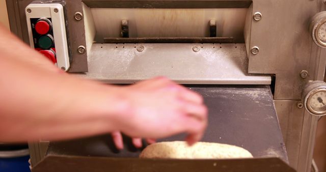 The photo features a baker's hand placing a loaf of dough into an industrial bread slicing machine. The image captures the precision and equipment used in a professional bakery or food production facility. This image can be ideal for illustrating bakery processes, commercial kitchen equipment, or industrial baking settings.