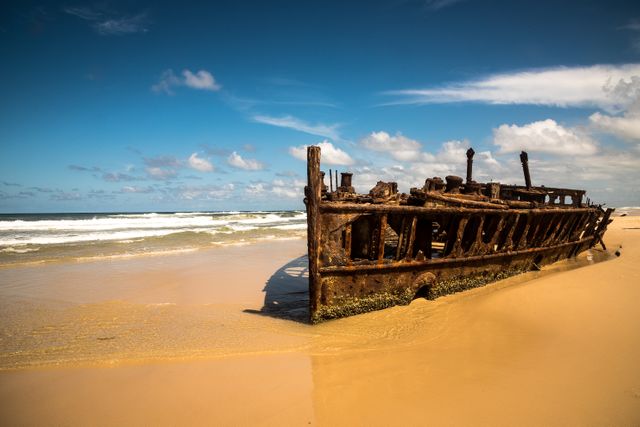Rusted shipwreck lying on sandy beach under clear blue sky with gentle ocean waves in the background. Ideal for use in travel brochures, maritime history articles, nature photography collections, and beach-themed decor. This evocative seaside scene captures the beauty of deterioration and the maritime history.