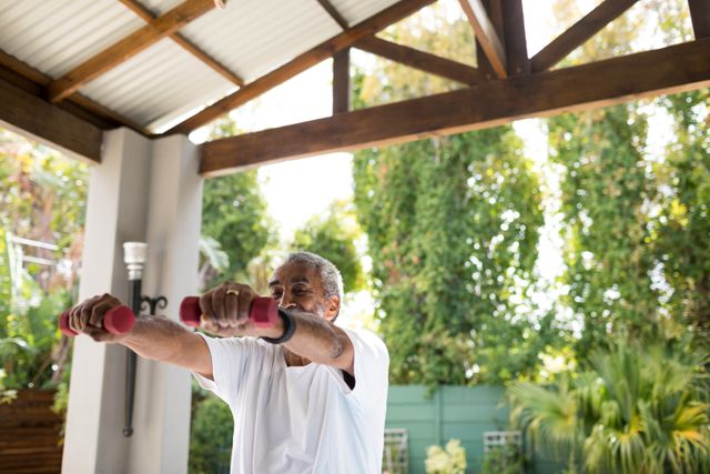 Senior man lifting dumbbells in backyard under shed roof. Suitable for use in health and fitness campaigns, wellness blogs, active aging advertisements, and articles promoting physical exercise for elderly.