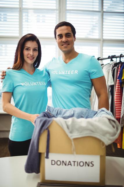 Happy volunteer couple standing with a donation box filled with clothes in a bright workshop. They are wearing matching 'Volunteer' T-shirts, smiling at the camera. Can be used for promoting volunteer work, charity events, community service projects, nonprofit organizations, and donation drives.