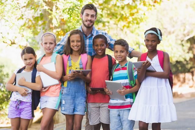 This image shows a teacher standing with a diverse group of school children outdoors. The children are holding tablets and wearing backpacks, indicating they are ready for school activities. The setting is bright and cheerful, with trees in the background. This image can be used for educational materials, school promotions, diversity and inclusion campaigns, and back-to-school advertisements.