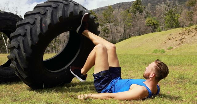A fit man in athletic wear is flipping a heavy tire in an outdoor setting. The environment is a grassy field with a background of trees and hills. This image is ideal for use in fitness, sports, and health-related advertising, promotional materials, and blog content.