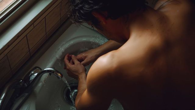 Shirtless man leaning over sink cupping water under chrome faucet in warm bathroom light