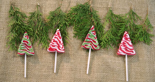 Four Christmas tree-shaped lollipops are arranged in a row against a burlap background with evergreen sprigs above them, with copy space. Festive and colorful, these candies evoke the cheerful spirit of the holiday season.
