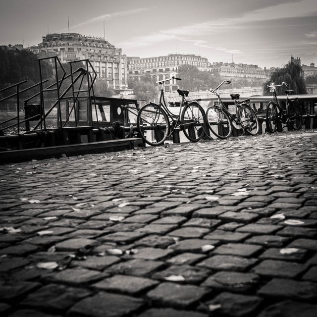 Bicycles are parked along a cobblestone street near a river with historic buildings in the background. Taken in black and white, this image evokes a vintage and nostalgic ambiance. Perfect for travel blogs, historical articles, or as a decorative piece highlighting European charm and transportation themes.