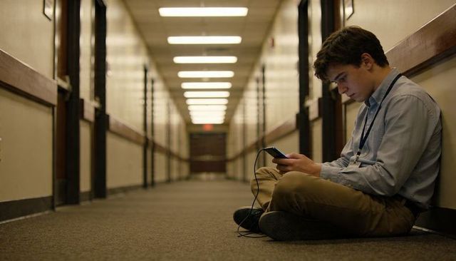 High school student sitting in corridor checking phone with charging cable and lanyard