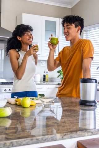 Multiracial couple enjoying green smoothies in bright modern kitchen, laughing together