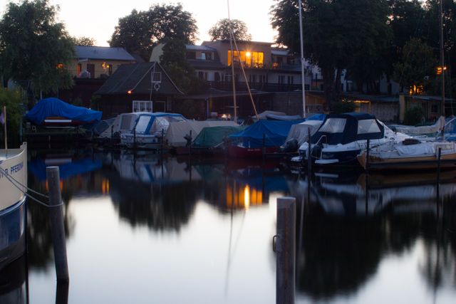Marina during evening twilight with boats covered and docked, surrounded by tranquil water reflecting the buildings and lights. Suitable for topics related to nature, tranquility, waterfront living, residential areas, boating, and relaxation.
