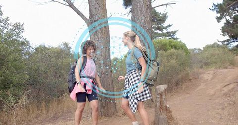 Two women walking and chatting on sunlit trail with backpacks and futuristic blue overlay