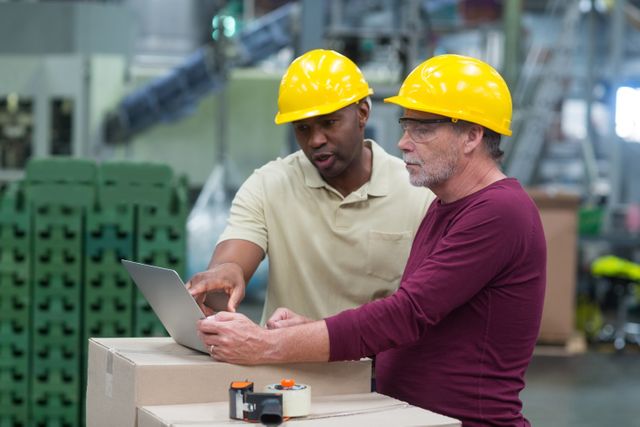 Two factory workers wearing hard hats are discussing production details while working on a laptop in a drinks production plant. This image can be used to illustrate teamwork, industrial processes, and the integration of technology in manufacturing environments. Ideal for articles or presentations on factory operations, workplace safety, and industrial efficiency.