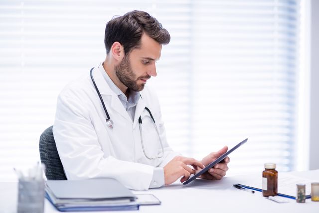 Young male doctor using digital tablet while sitting at desk. Technology integrating in medical practice. Ideal for healthcare, technology presentations, medical blogs, telemedicine concepts, and promotional materials highlighting modern healthcare professional environments.