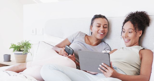 Two young women lounging on bed using tablet and smartphone, smiling together - Download Free Stock Images Pikwizard.com