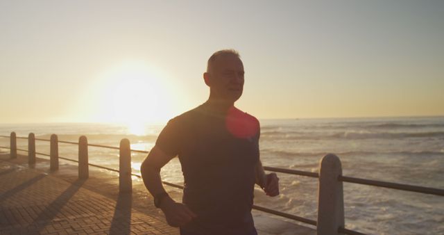 Mature man jogging along a seaside walkway at sunrise, promoting a healthy and active lifestyle. Great for use in articles or advertisements related to fitness, health and wellness, senior activities, and outdoor exercise.