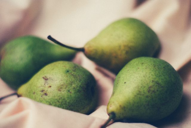 Close-up shot of four green pears resting on a soft, light-colored cloth, with natural light highlighting their fresh, ripe appearance. Ideal for use in culinary blogs, healthy eating articles, and organic produce promotions.