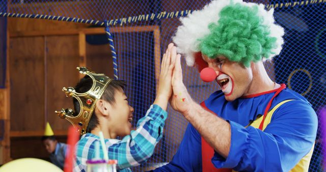 A young Asian boy is giving a high-five to a clown at a festive event, with copy space. Their joyful interaction captures the lively atmosphere of a child's birthday party or a family-friendly celebration.