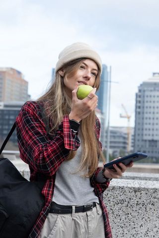 Female commuter wearing cream beanie leaning on railing holding green apple and phone