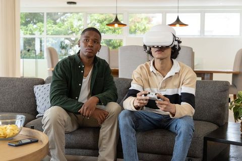 Two diverse friends playing virtual reality game on sofa while one wears VR headset and holds contro