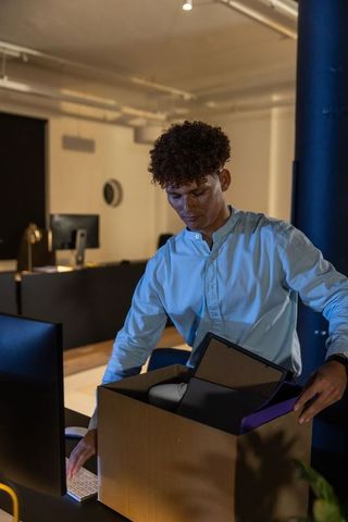 African american man packing office supplies into cardboard box at desk working late