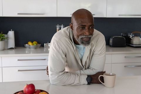 Mature african man leaning on modern white kitchen countertop holding mug, reflecting