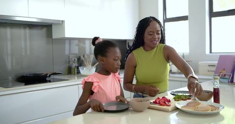 Mother and daughter preparing pancakes at kitchen island adding strawberries and syrup