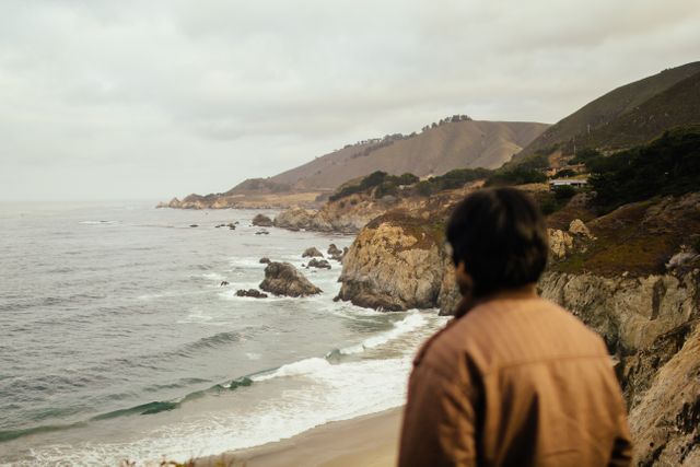 Man observing rugged coastline and rolling waves on a cloudy day. Great for depicting solitude, contemplation, connection with nature, and peaceful coastal scenes. Suitable for travel blogs, nature articles, mental health content, and outdoor adventure marketing.