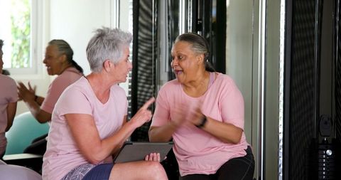 Senior multiracial lesbian couple in gym joyfully using tablet