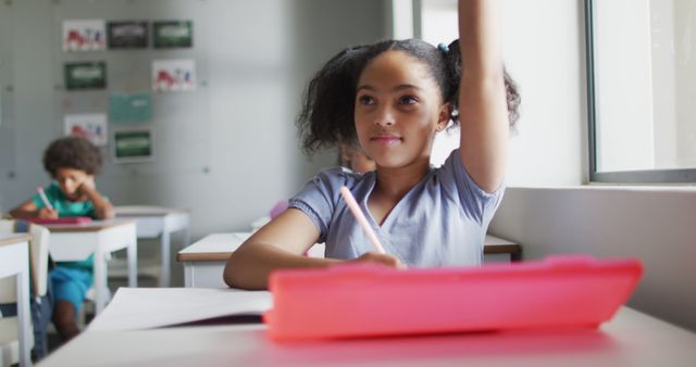 This captures a young girl enthusiastically raising her hand during a class, seated at her desk with a bright folder open in front of her. She is focused and ready to participate in the lesson. This is ideal for educational materials, school brochures, academic websites, or content highlighting student engagement and active learning.