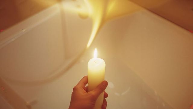 Hand Holding Lit Candle Above Bathtub in Dimly Lit Bathroom