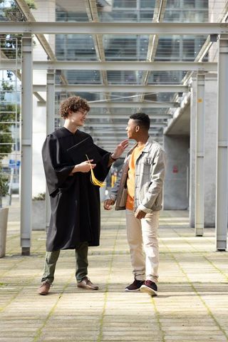 Multiracial graduate wearing gown chatting with friend on campus walkway under canopy